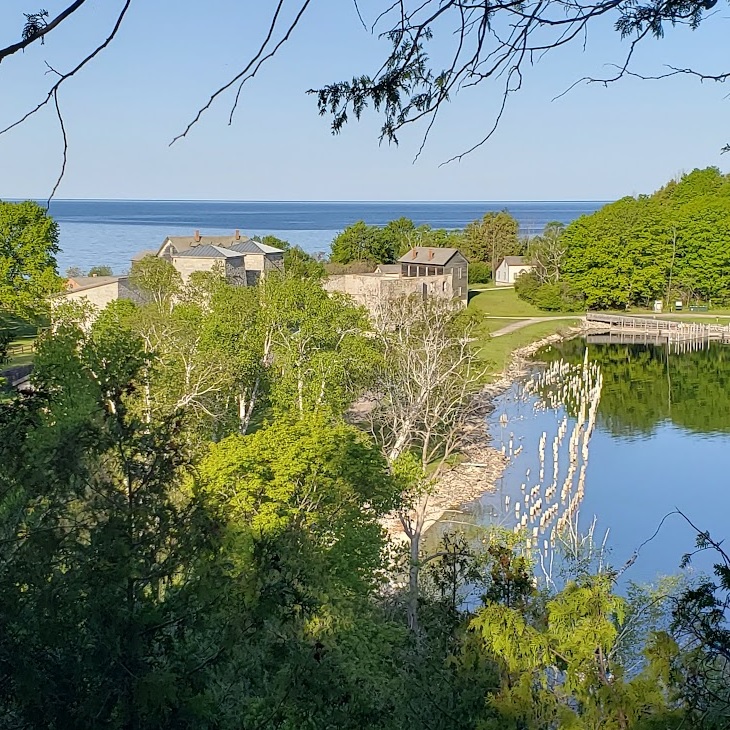 The ghost town of Fayette in the near distance seen from the edge of the limestone cliffs. Visible are the furnace stacks, the company store, the town hall, a single-family house, and the roof of the hotel. The pilings of the old wharf jut from the surface of the water in the harbor. Leafy green trees frame the photo. 