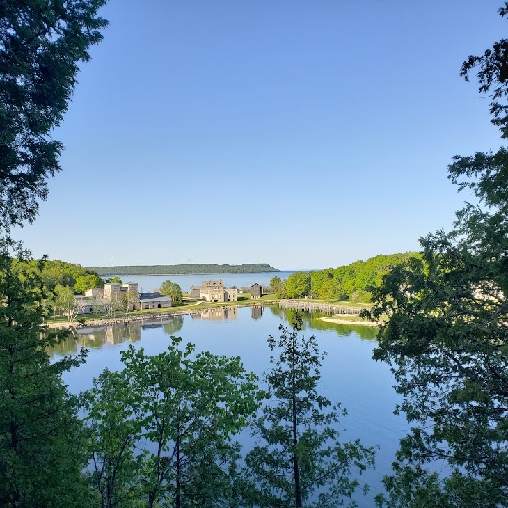 The ghost town of Fayette in the middle distance seen from the edge of the limestone cliffs. Visible are the furnace stacks, the casting rooms, the company office, the roof of the hotel,  the company store, and the town hall. A peninsula covered in trees is visible in the background. Leafy green trees frame the photo. 