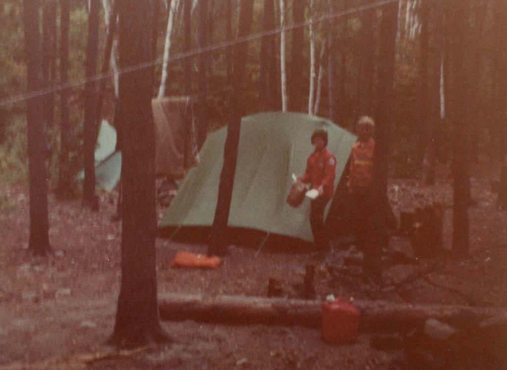 two boys wearing brightly colored jackets stand in front of a green tent, surrounded by tall trees