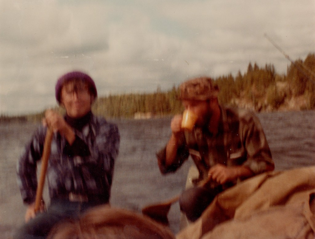 blurry photo of two people in flannel shirts sitting in canoes; one is paddling, one is drinking from a tan mug; the water is choppy and the sky is partly cloudy