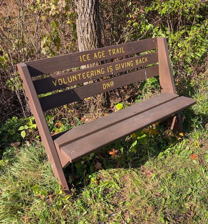 a brown wooden bench in the woods; the words "Ice Age Trail - Volunteering is giving back - DNR" are carved into the backrest