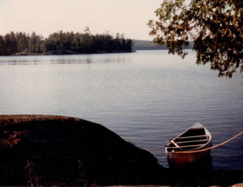 A calm lake with partly cloudy skies overhead; a canoe is tied up at the shore in the foreground; trees crowd the shoreline in the background