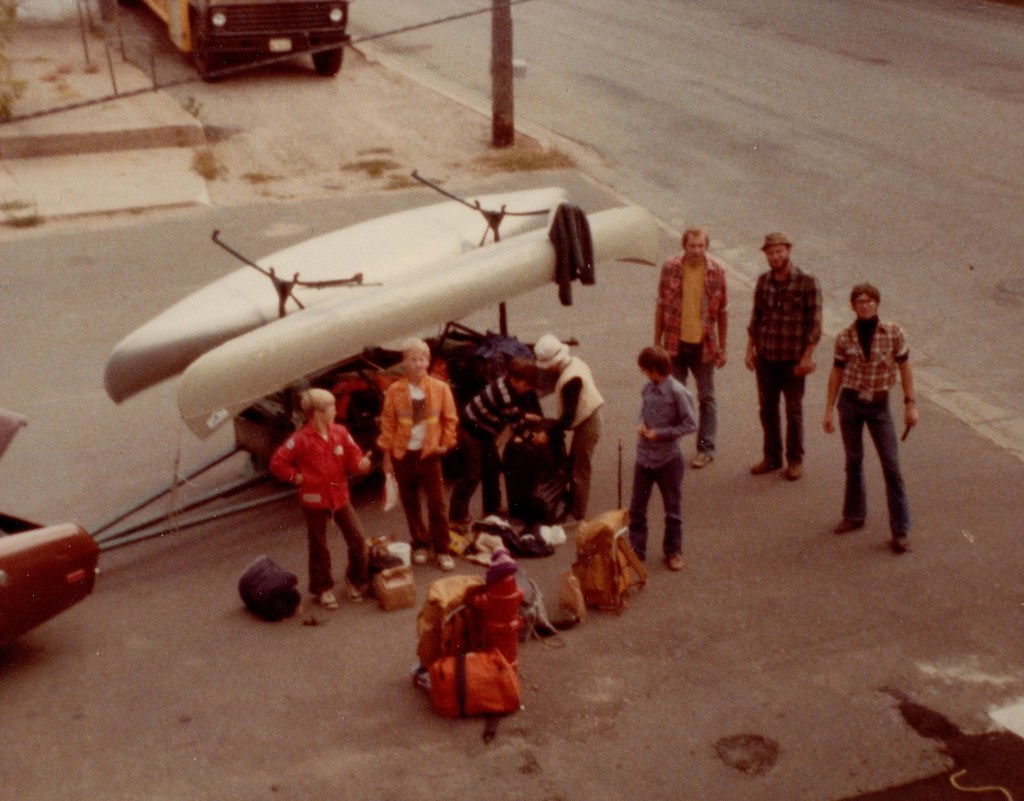 eight people stand in front of a canoe trailer; two canoes are loaded onto the racks and the bottom of the trailer is packed full of camping gear