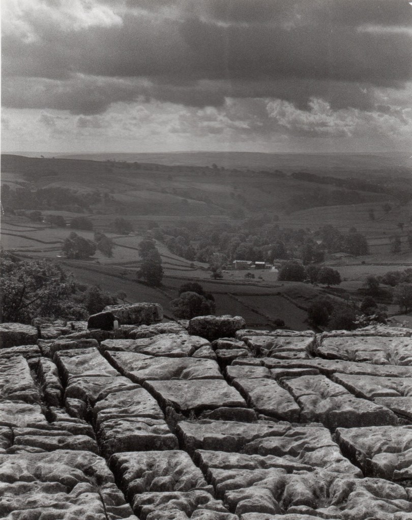 the foreground is limestone, pitted and cracked; farm fields dotted with leafy trees stretch away into the far background, many hundreds of feet below; the sky is broken clouds the color of iron