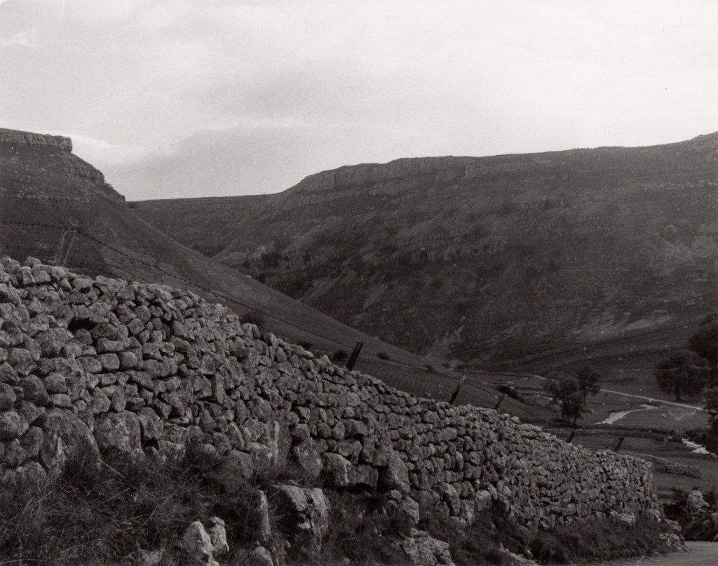 a dry stone wall stretches across the foreground; a steep slope falls from left to right in the middle ground; a very steep cliff looms in the background; the sky is overcast