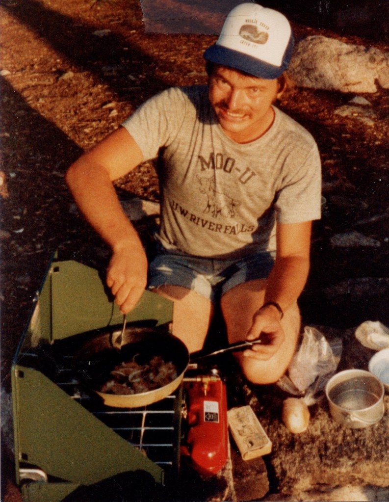 a young man kneels over a Coleman gas stove, where he is pan-frying the perch he caught earlier in the evening