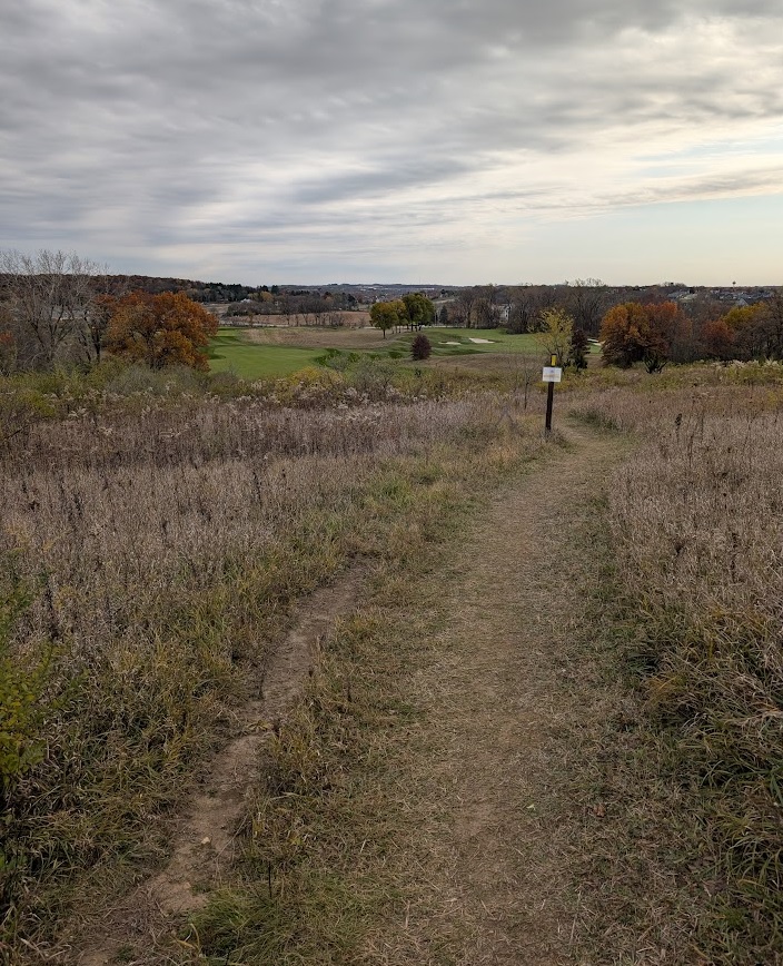 a hiking trail through short field grass in the foreground; manicured golf links in the background; a cloudy sky overhead