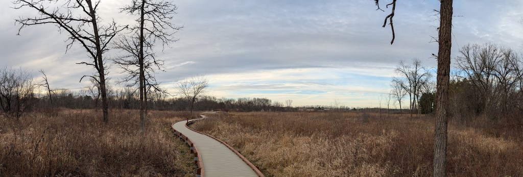 a boardwalk across a wetland in the foreground; bare trees border the horizon in the background; an overcast sky hanged over the scene