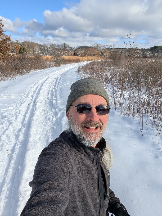 man with a short beard and dark glasses wearing a wool beanie cap and a grey jacket stands on a snow-covered field; the sky is partly obscured by puffy gray clouds