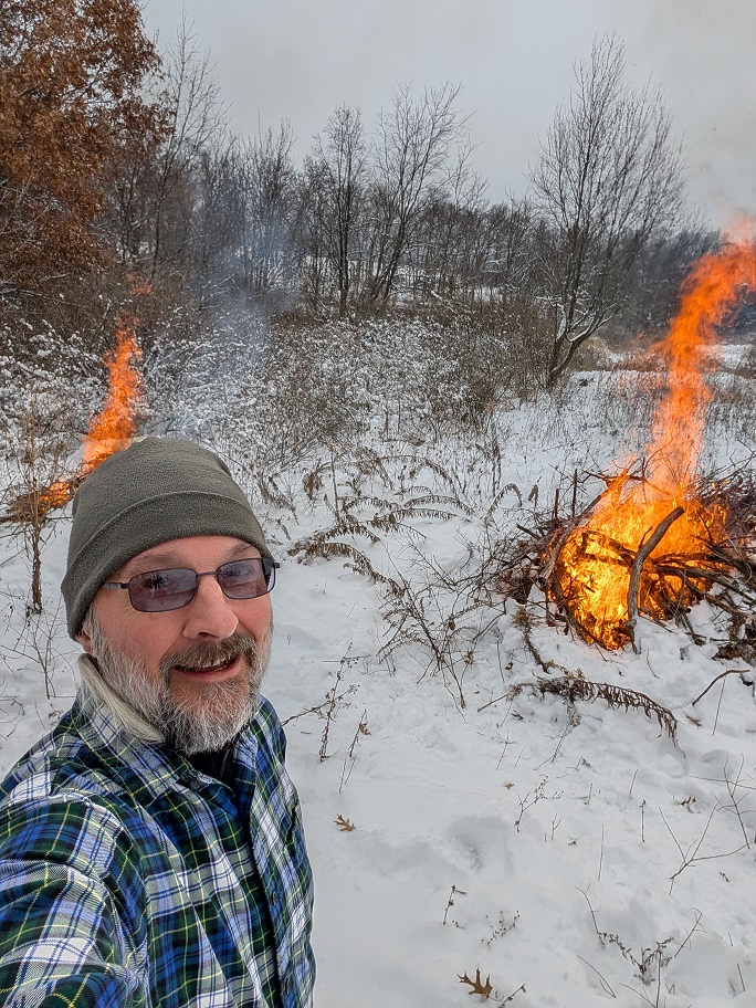 a man in a green beanie cap and blue checked flannel shirt appears to be very happy that piles of cut buckthorn and honeysuckle are going up in flames behind him