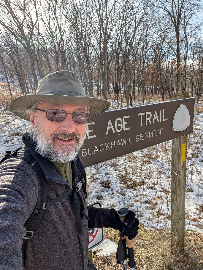 the author, yours truly, standing in front of a sign reading "Ice Age Trail Blackhawk Segment" - I am wearing a wide-brimmed hat, dark glasses, a grey jacket over a green shirt, and I'm holding a pair of trekking poles