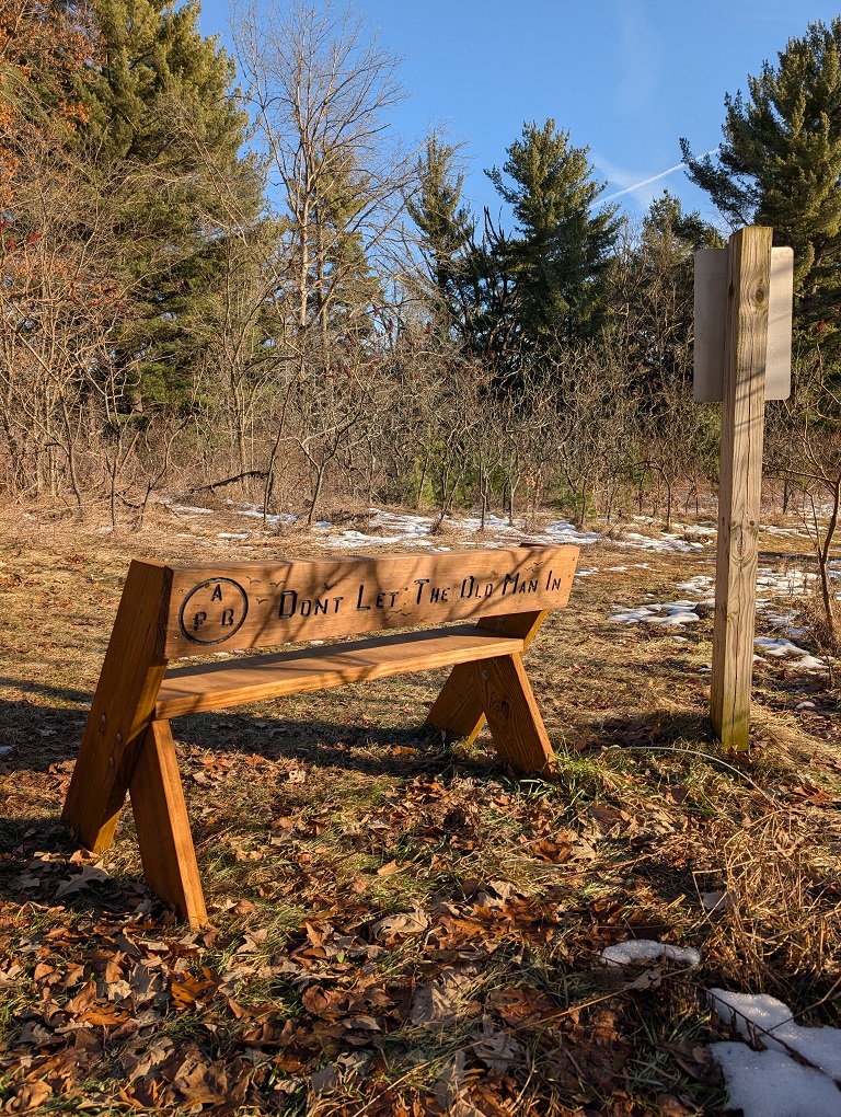 a Leopold bench bearing the epithet "Don't Let The Old Man In" stands alongside a trail through a forest