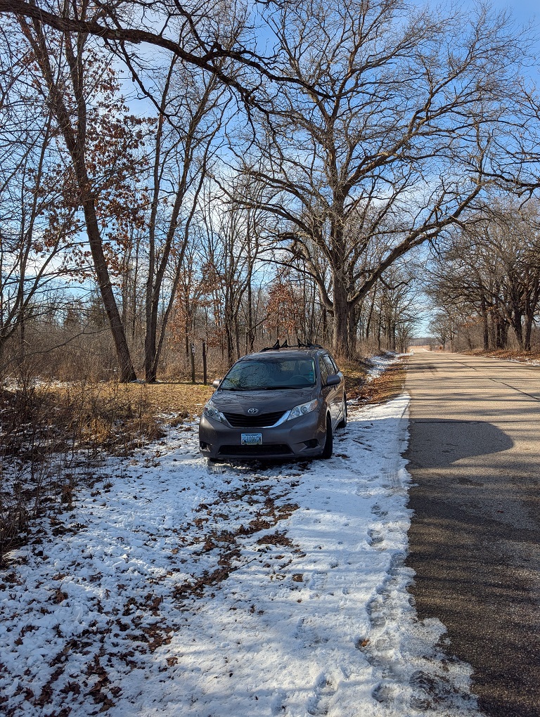 a dark gray minivan is parked on the snow-covered shoulder of a country road; tall trees tower overhead; the sky is mostly clear with high wispy clouds