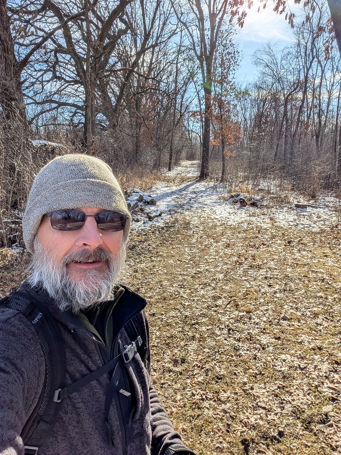 a bearded man in dark glasses poses alongside a wooded trail; he is wearing a gray wool beanie cap and a dark gray jacket