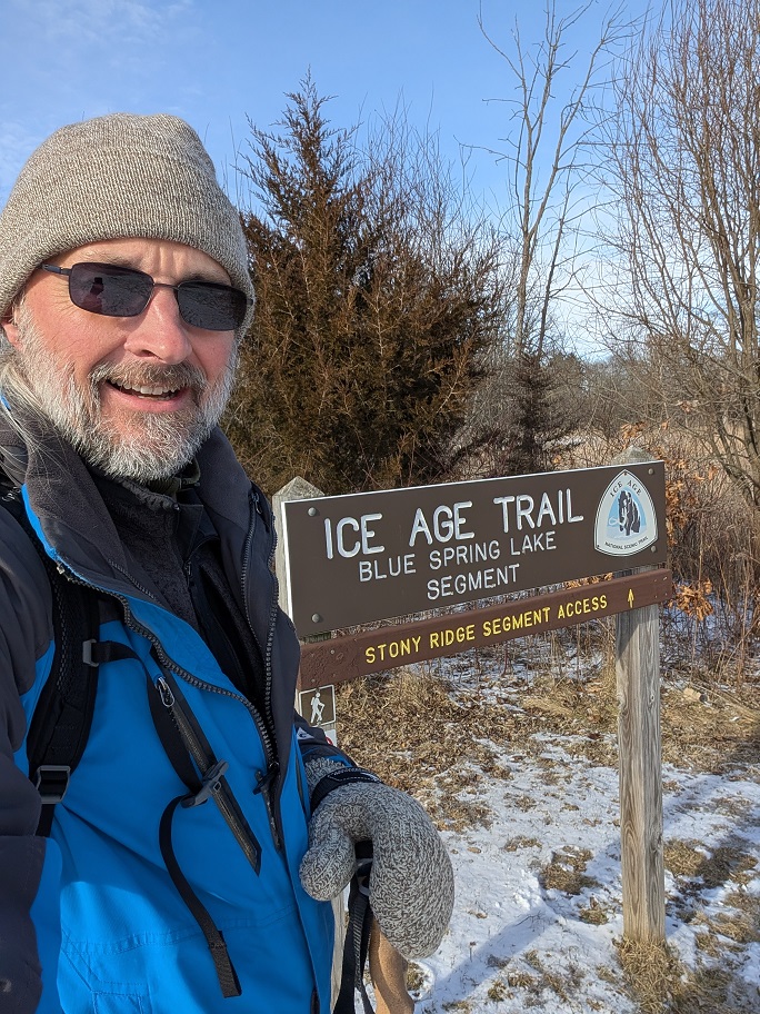 a bearded man in dark glasses poses next to a trail sign; he wears a gray wool beanie cap, a blue overcoat and thick woolen mittens