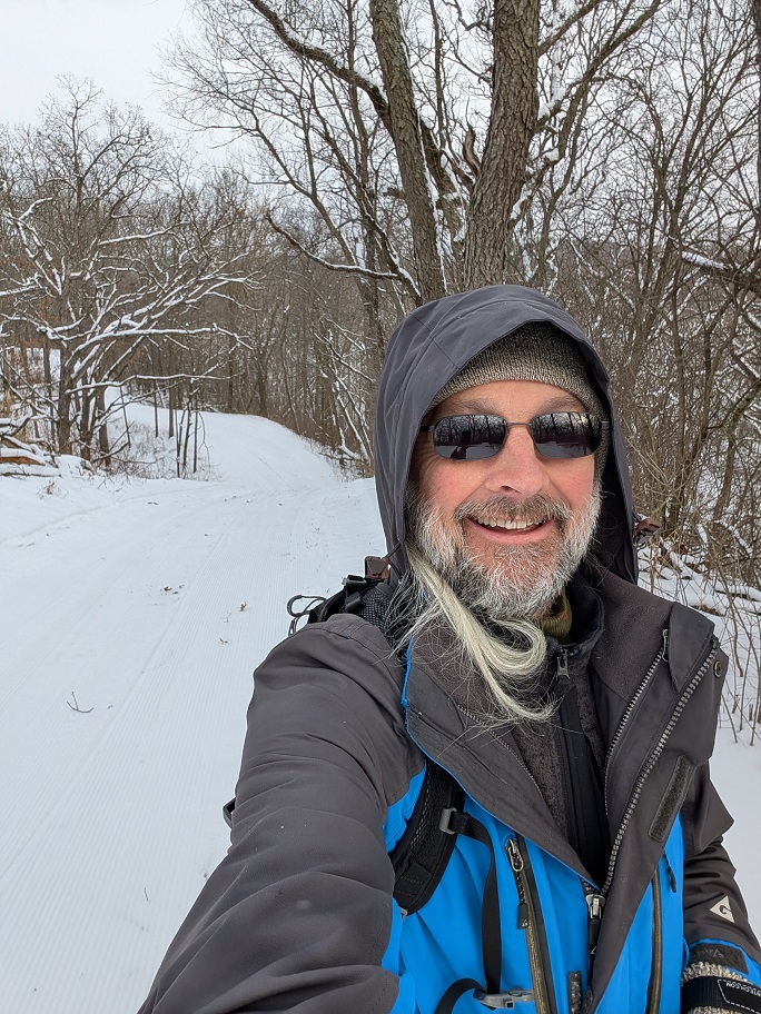 a bearded man in a dark hooded overcoat and dark glasses poses alongside a snow-covered trail through a wooded area