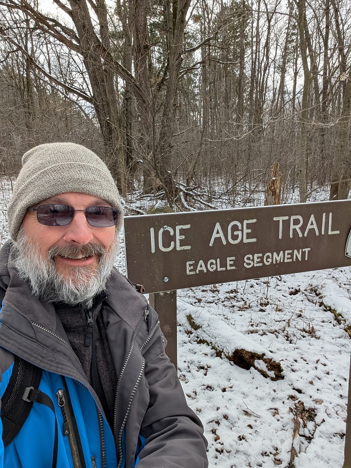 a bearded man in dark glasses poses in front of a sign which reads "ICE AGE TRAIL - EAGLE SEGMENT"
