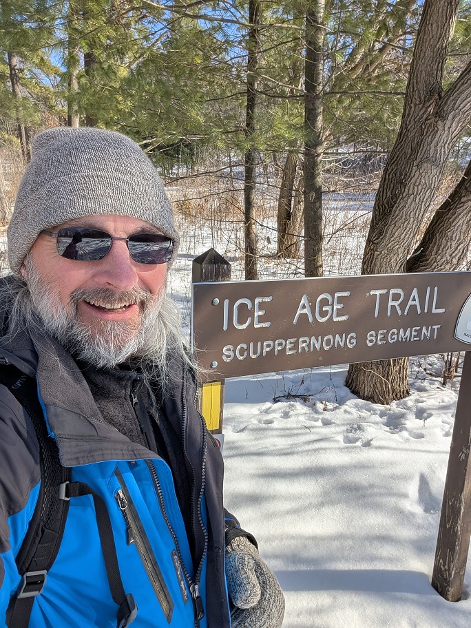 a bearded man in a gray wool cap and blue nylon overcoat poses beside a trailhead sign