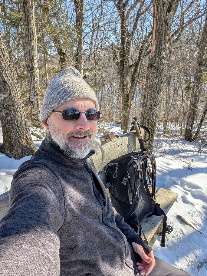a man with a gray beard poses on a bench to take a selfie; he is wearing a gray beanie cap and a charcoal-colored fleece jacket