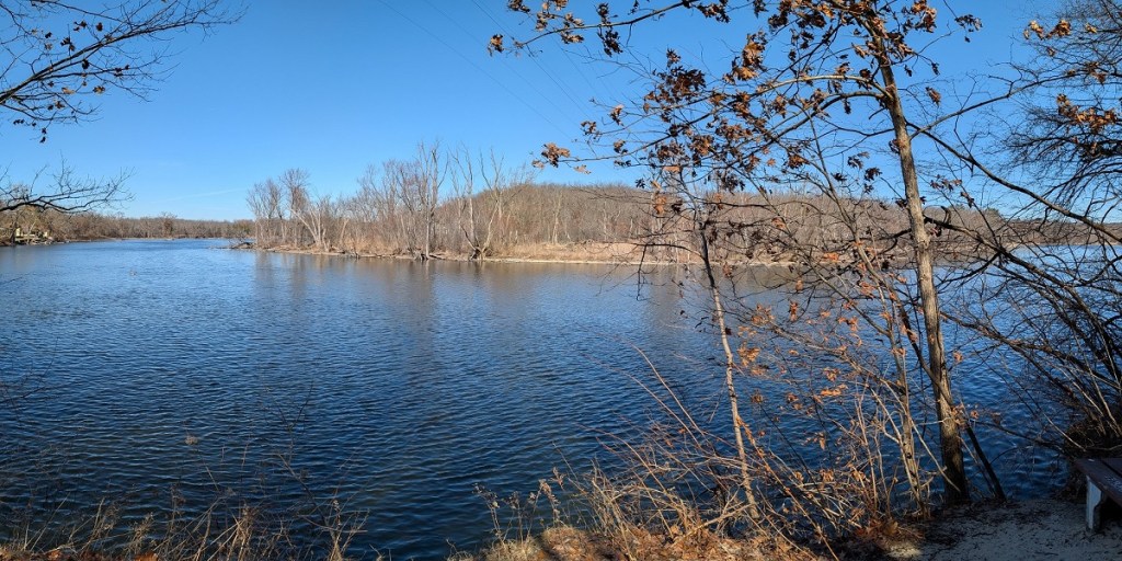 a calm river flowing through a winter landscape; the trees are almost entirely leafless though a few brown leaves hang on; the sky is a brilliant blue