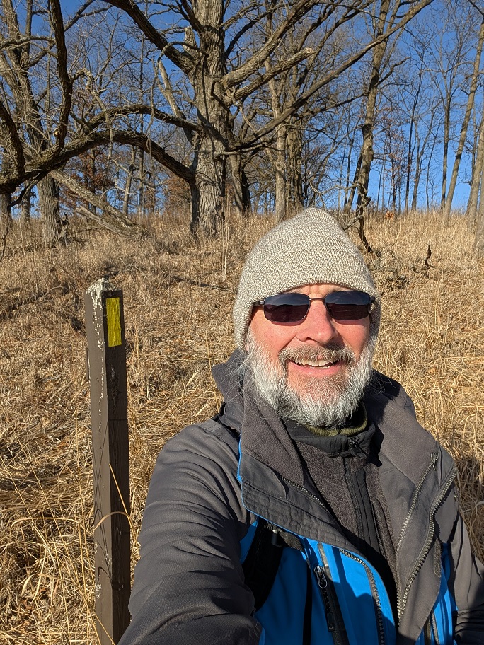 a bearded man in a gray beanie cap and dark glasses poses in a field of dry brown prairie grass; leafless oak trees tower over his head in the background